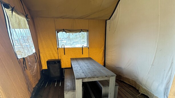 A tent with a wooden table and bench, a window, and a trash bin.
