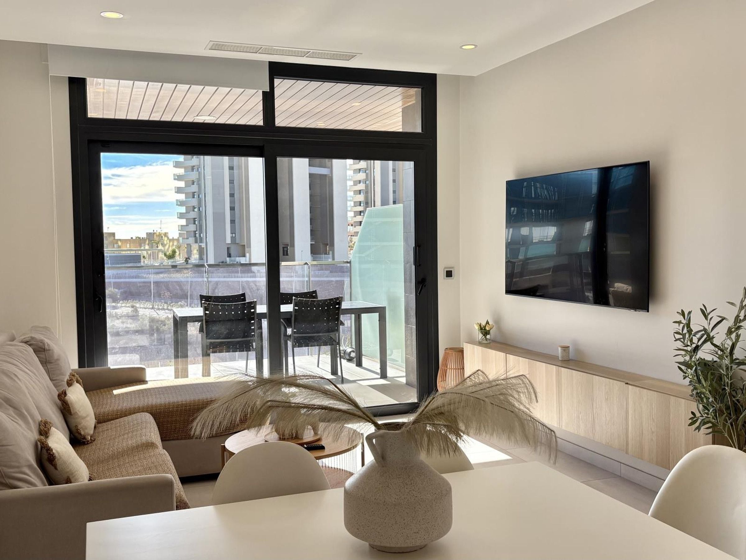 A modern living room with a sofa, a dining table, and chairs. There is a large window with a view of buildings outside.