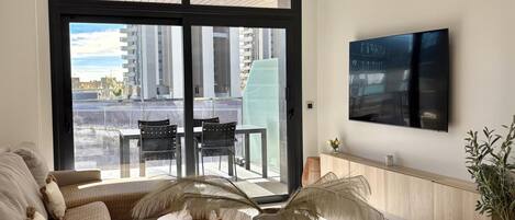 A modern living room with a sofa, a dining table, and chairs. There is a large window with a view of buildings outside.