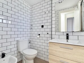 Ein Badezimmer mit weißen Fliesen, einer weißen Toilette, einem Waschbecken mit Spiegel und einer Holzkommode.