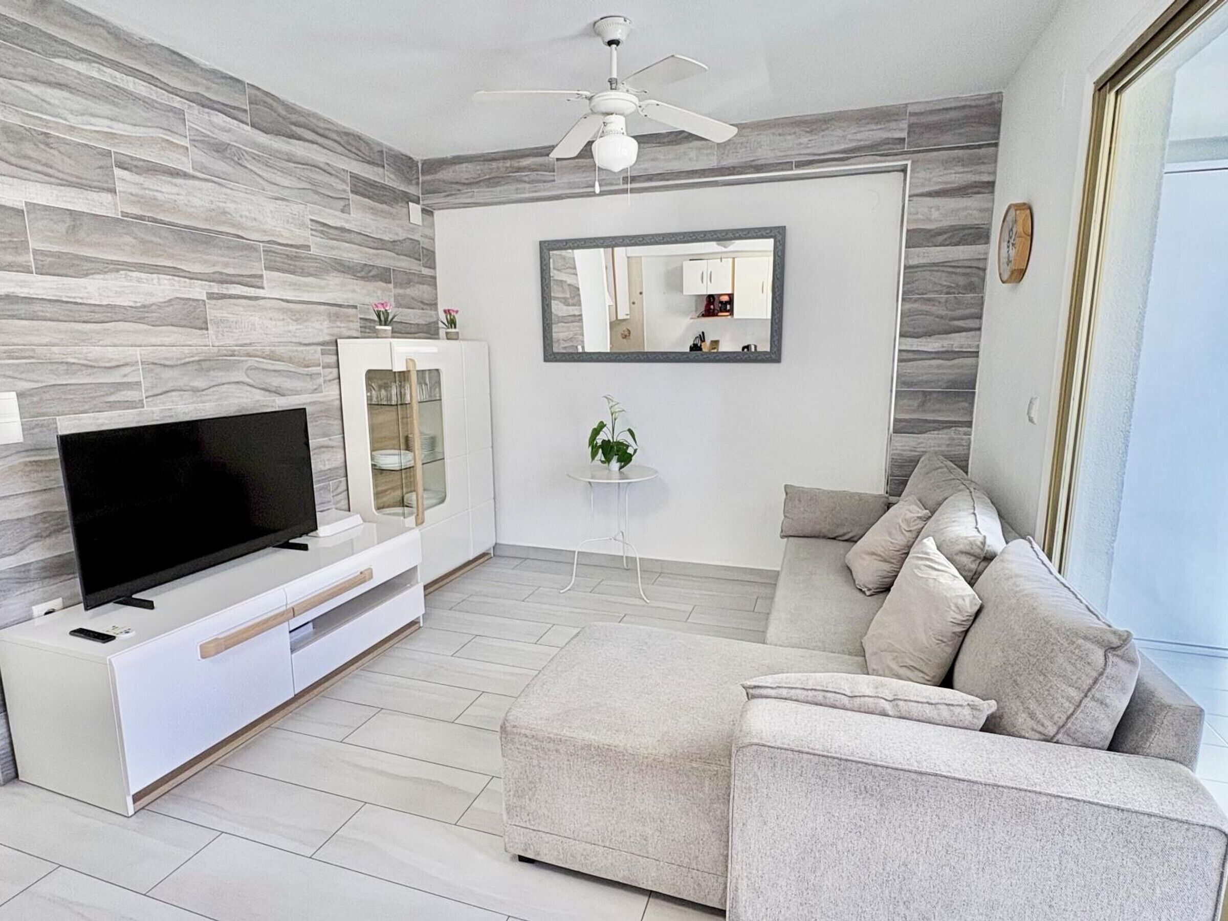 Een moderne woonkamer met een flatscreen televisie, een witte entertainmentkast, een grijze bank en een plafondventilator.