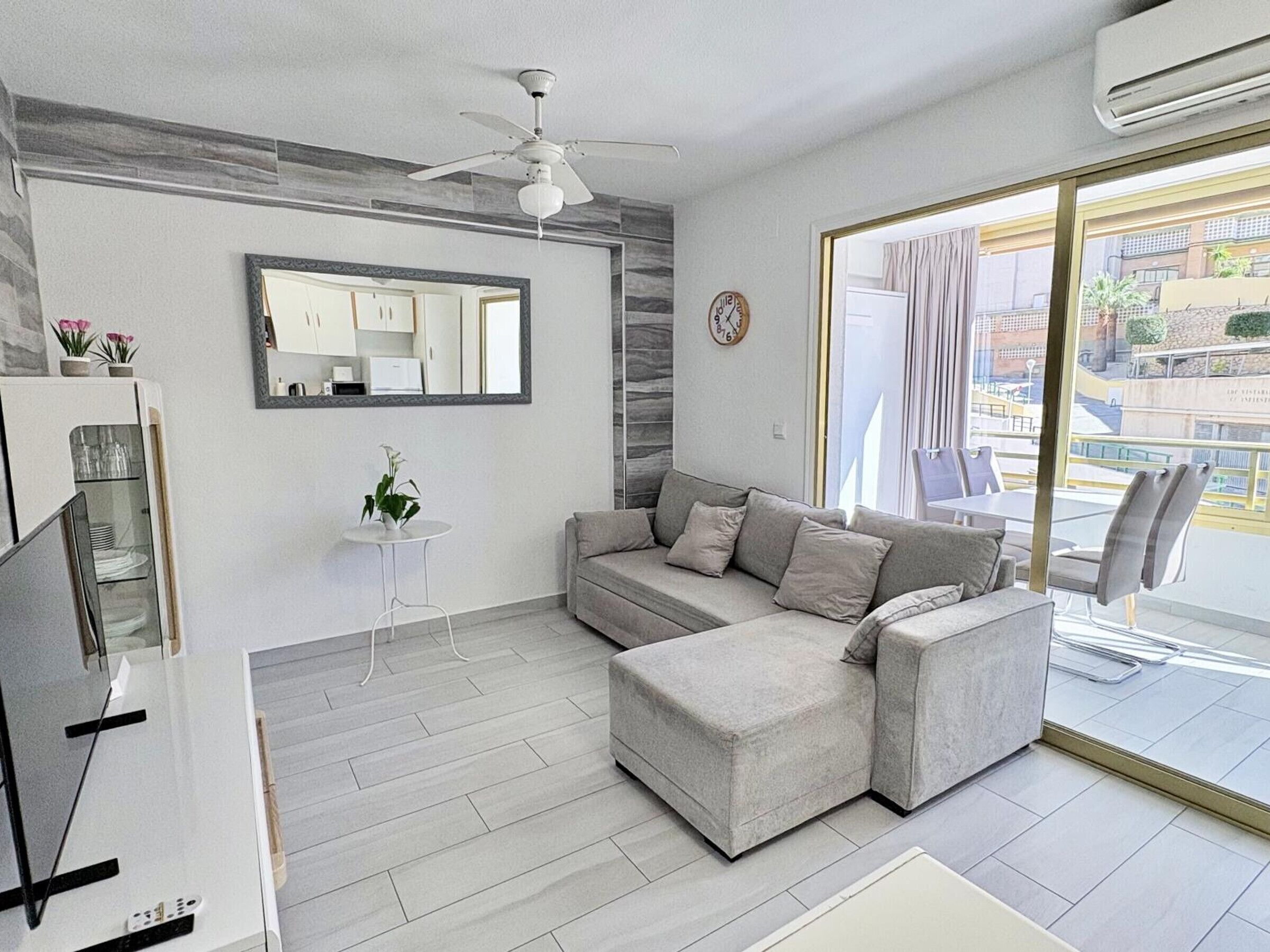 A modern living room with a grey sofa, a small white side table, a ceiling fan, and a large window with a view of the outside.
