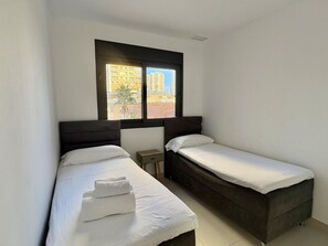 Ein Zimmer mit zwei Betten, einem Fenster mit Blick auf Gebäude und einem kleinen Tisch.