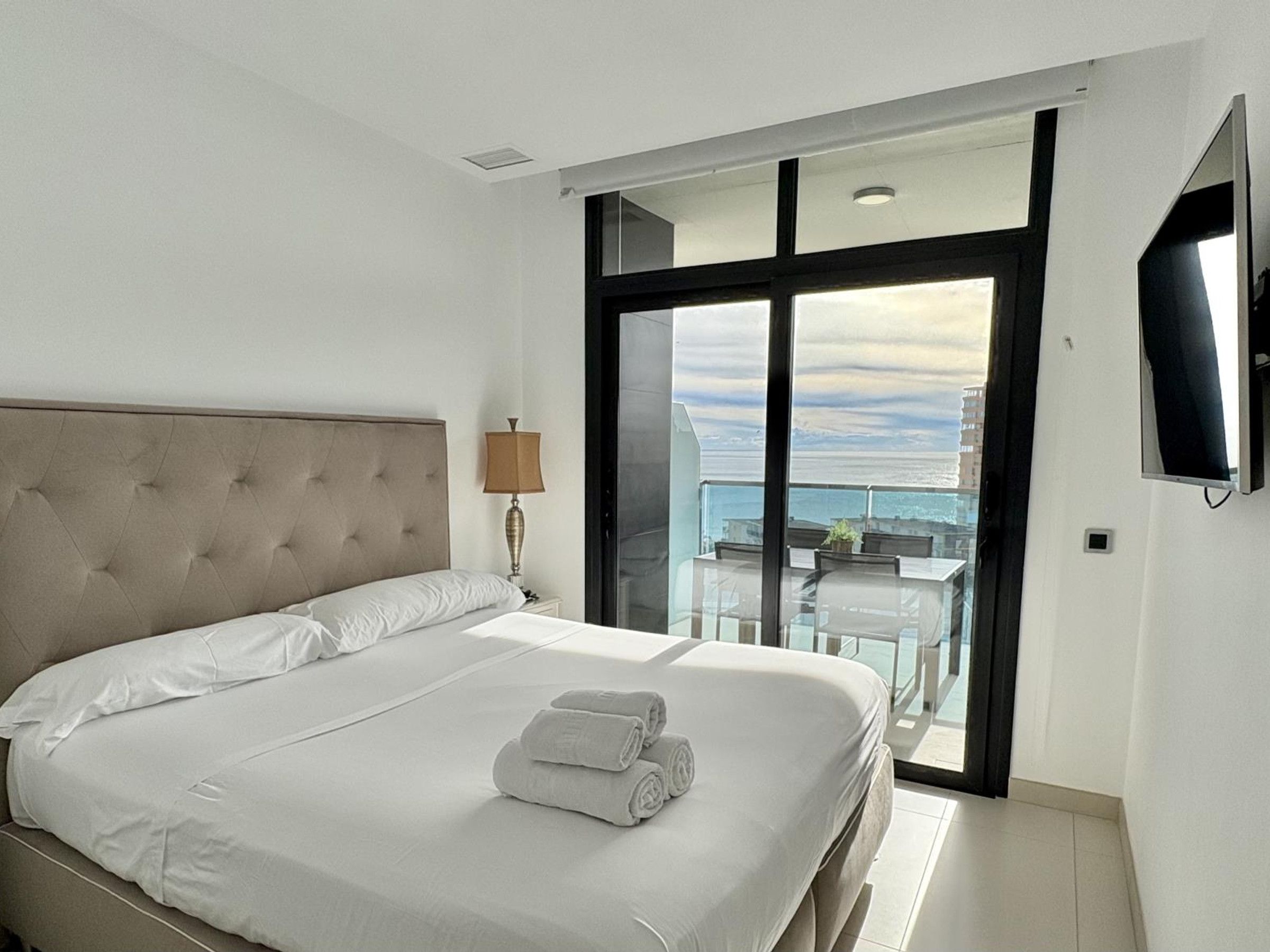 A hotel room with a large bed, a bedside lamp, a TV, and a balcony with a table and chairs.