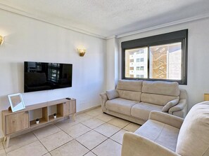 Un saló amb televisió de pantalla plana, un sofà beix, un moble de roba de fusta i una finestra amb vista a edificis.
