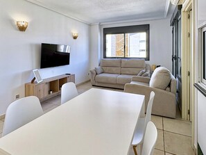 A modern living room with a flat-screen TV, a beige sofa, a dining table with chairs, and a window with a view of buildings. - Modern apartment near the beach - 4 guests (Villajoyosa)