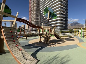 Ein Spielplatz mit Rutsche und Klettergerüsten, umgeben von einem befestigten Bereich, mit einem Berg im Hintergrund.