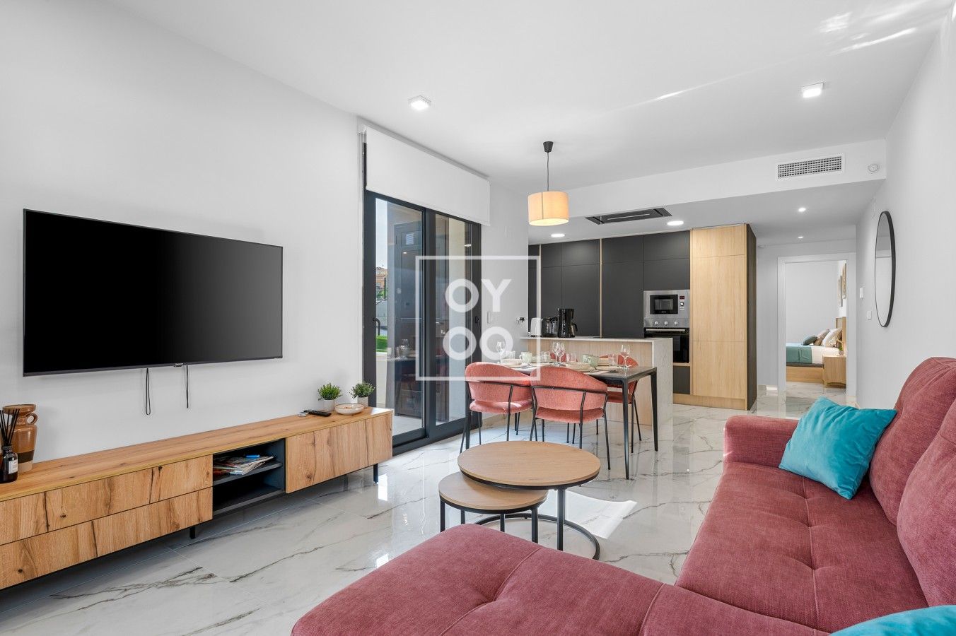 Una sala moderna con un televisor de pantalla plana, un mueble bajo de madera, un sofá rojo y un comedor con sillas y una mesa.