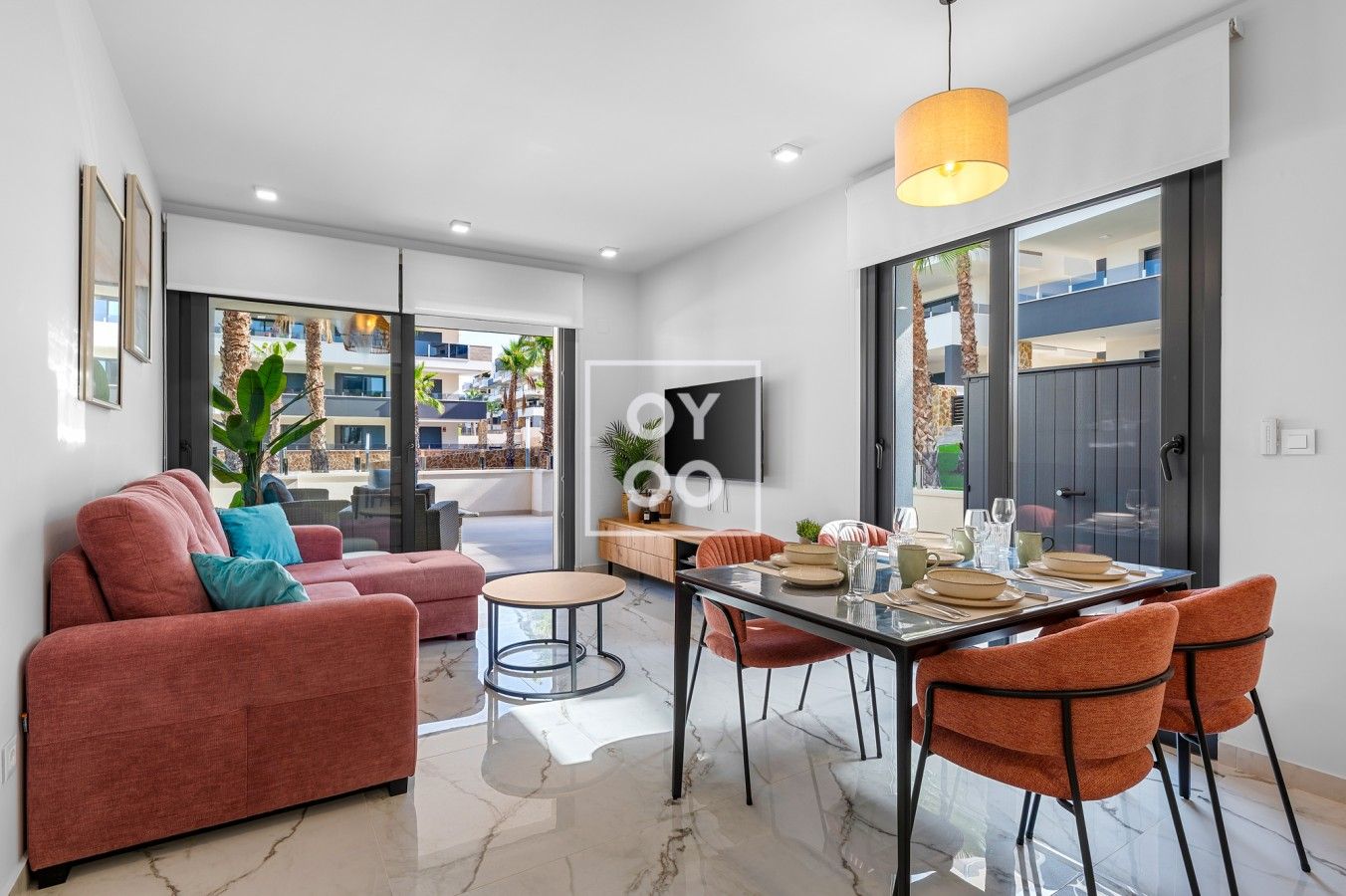 A modern living room with a sofa, a dining table with chairs, a TV, and a large window with a view of palm trees.