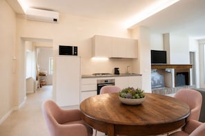 A modern kitchen with white cabinets, a microwave, and a stove. There is a dining area with a wooden table and pink chairs. - Vrbo Property (arezzo)