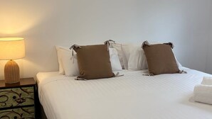 A neatly made bed with white bedding and brown pillows, a bedside lamp, and a decorative nightstand with a woven design. - Wagga Apartments #10 (Wagga Wagga)
