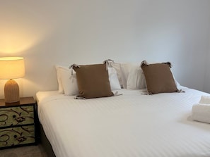 Een net opgemaakt bed met witte lakens en bruine kussens, een bedlampje en een decoratief nachtkastje met geweven details.