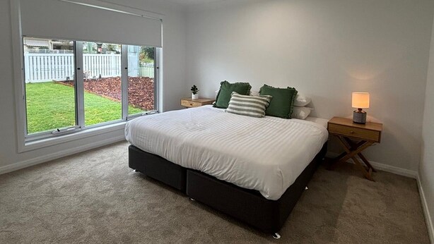 A bedroom with a large bed, a nightstand, a lamp, and a sliding glass door leading to a garden. - Wagga Apartments #10 (Wagga Wagga)