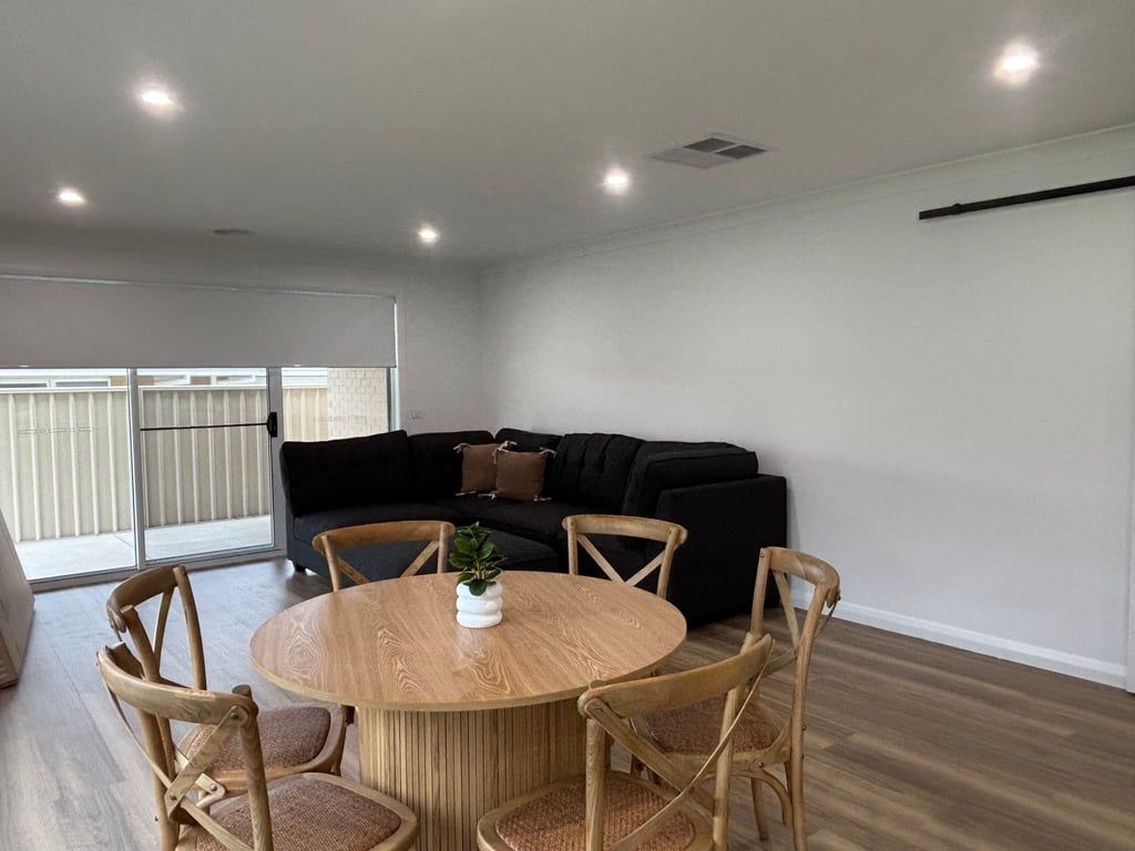 A modern living room with a round wooden table and chairs, a black sofa, and a large window with blinds.