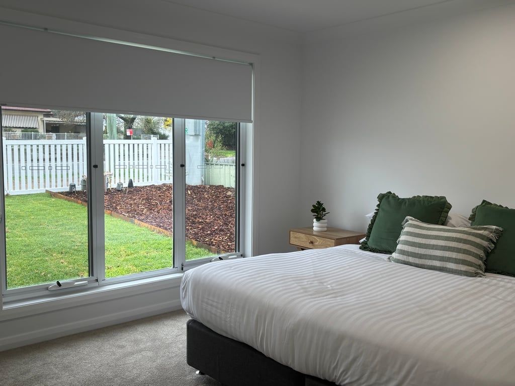 A bedroom with a large window, a bed with white bedding, a small bedside table with a plant, and two green pillows.