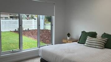 A bedroom with a large window, a bed with white bedding, a small bedside table with a plant, and two green pillows.