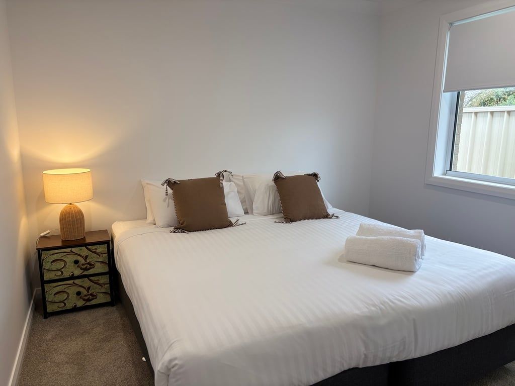 A neatly made bed with white bedding and two brown pillows, a bedside lamp on a nightstand with a decorative pattern, and a window with a view of a fence.