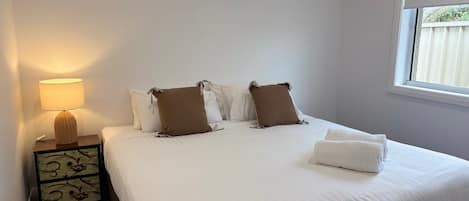Una cama bien hecha con ropa blanca y dos almohadas marrones, una lámpara de noche en una mesita con un motivo decorativo, y una ventana con vistas a una valla.