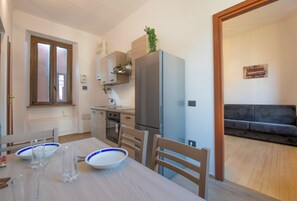 Une cuisine moderne équipée d’un réfrigérateur, d’un four et d’un évier. On y trouve une table à manger avec des chaises et un verre d’eau. Un canapé gris est visible au fond.