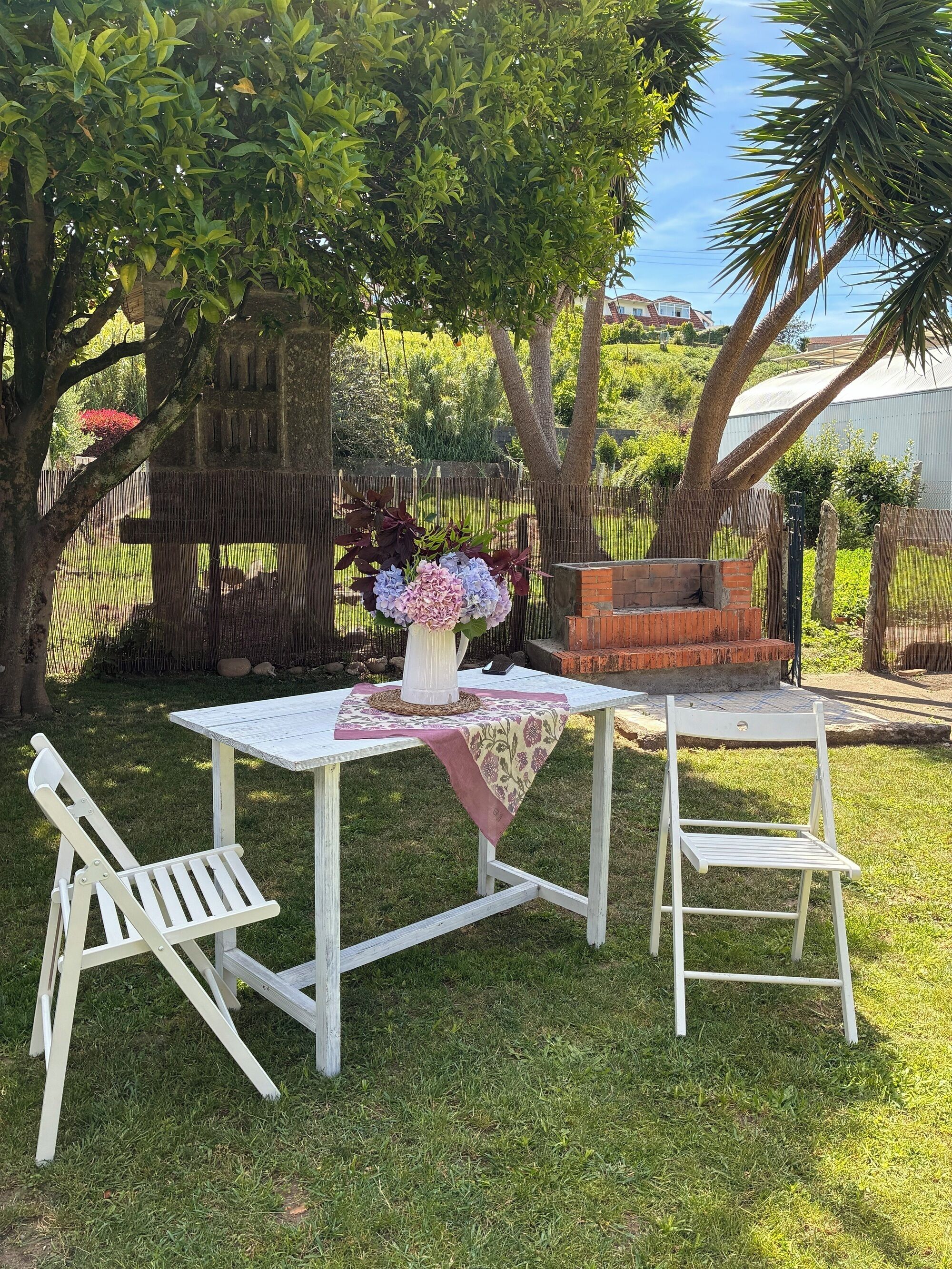 A white outdoor table with a floral centerpiece, two white chairs, and a brick barbecue grill in a grassy backyard with trees.