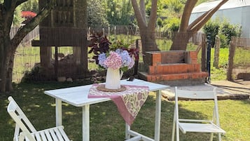A white outdoor table with a floral centerpiece, two white chairs, and a brick barbecue grill in a grassy backyard with trees.