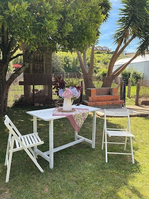 Une table blanche pour l’extérieur, agrémentée d’un arrangement floral, deux chaises blanches et un barbecue en briques, le tout installé dans une cour arrière verdoyante, parsemée d’arbres.