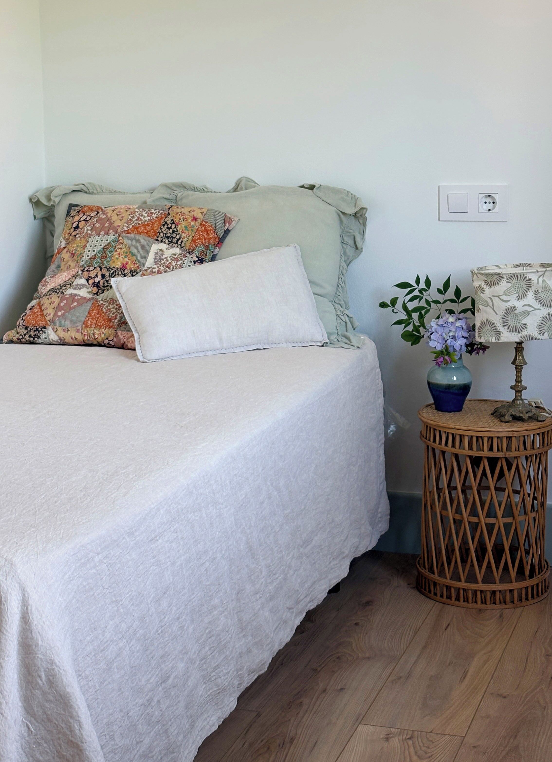 A bed with a white bedspread, a colorful quilted pillow, and a plain pillow. A small round table with a lamp and a vase of flowers is beside the bed.