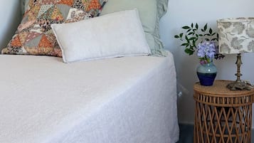 A bed with a white bedspread, a colorful quilted pillow, and a plain pillow. A small round table with a lamp and a vase of flowers is beside the bed.