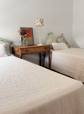 Un dormitorio con dos camas, una mesita de noche de madera con lámpara, un cuadro enmarcado y almohadones decorativos.