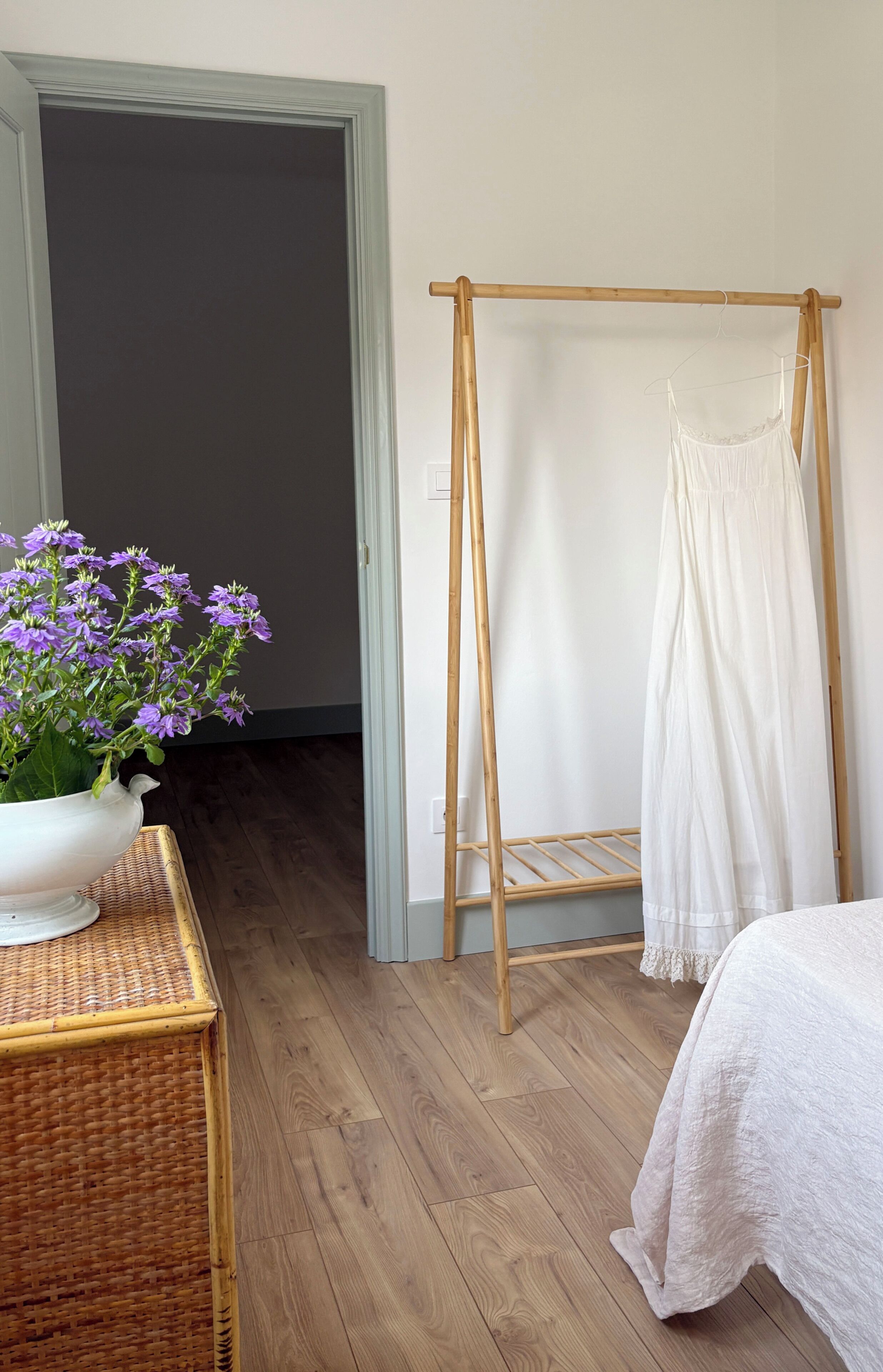 Een kamer met een houten vloer, een wit bed met een witte dekbedovertrek, een houten droogrek met een kledingstuk en een witte vaas met paarse bloemen op een rieten tafeltje.