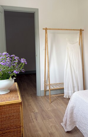 Um quarto com piso de madeira, uma cama branca com colcha branca, um varal de madeira com uma peça de roupa branca e um vaso branco com flores roxas sobre uma mesa de vime.