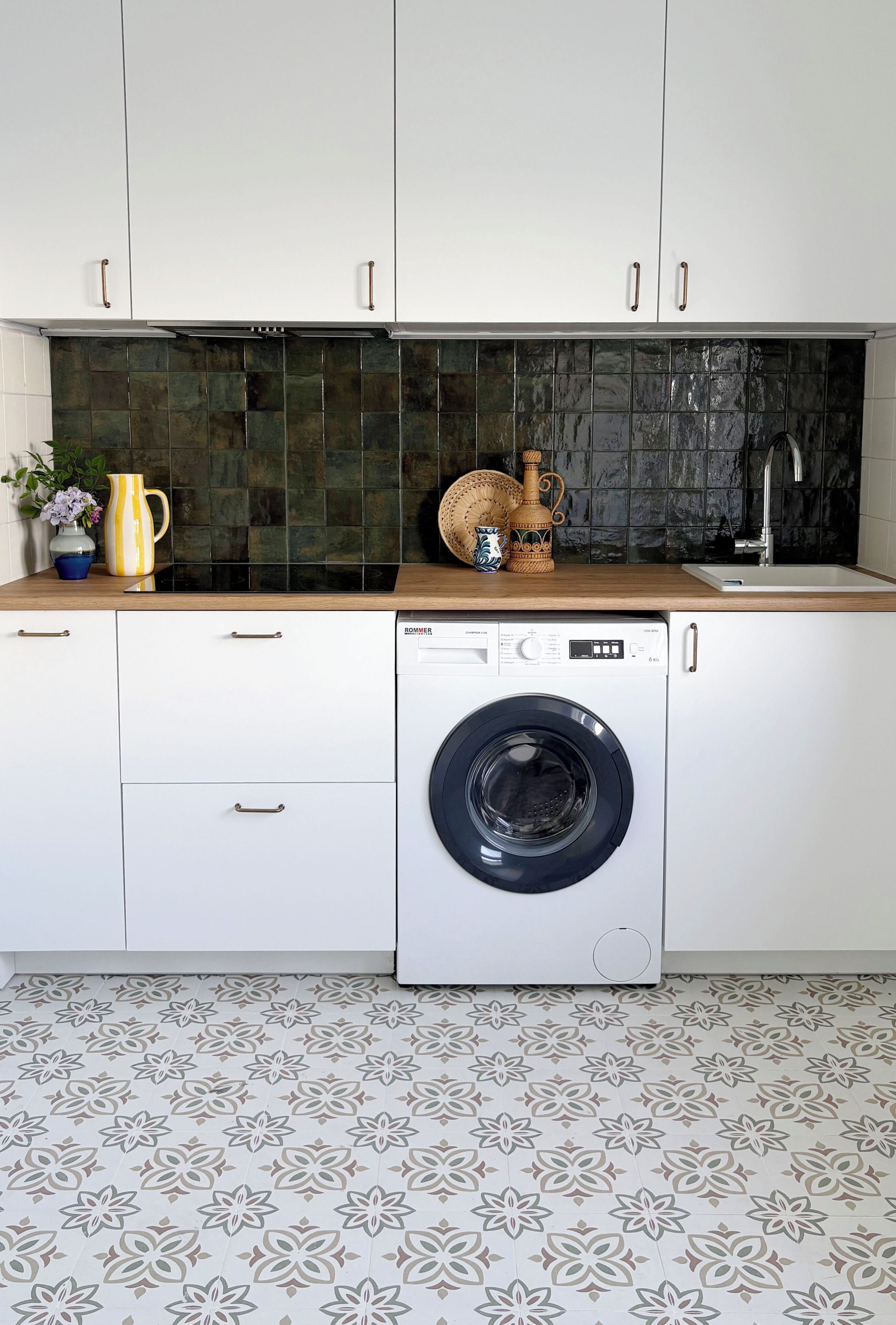 A compact kitchen with a front-loading washing machine, white cabinets, a dark backsplash, and a patterned floor.