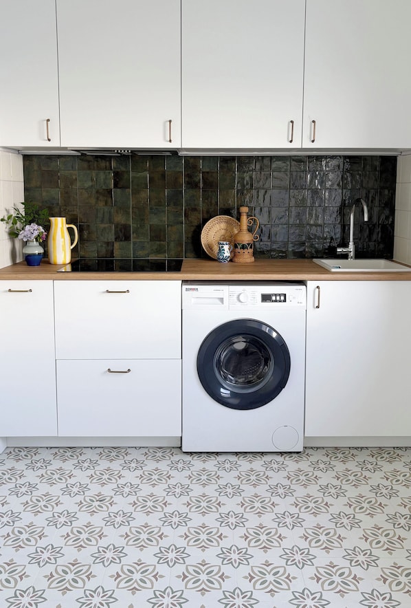 Une cuisine compacte dotée d’un lave-linge à chargement frontal, de meubles blancs, d’un plan de travail foncé et d’un sol à motifs.