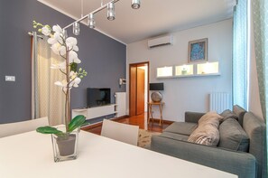 A modern living room with a grey sofa, a dining table with a potted plant, and a TV in the background. - Kedimore - Il Cuscino di Giulietta 2 (Verona)