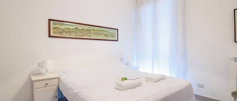 A hotel room with a bed, a nightstand, a lamp, a framed map on the wall, and a window with curtains.