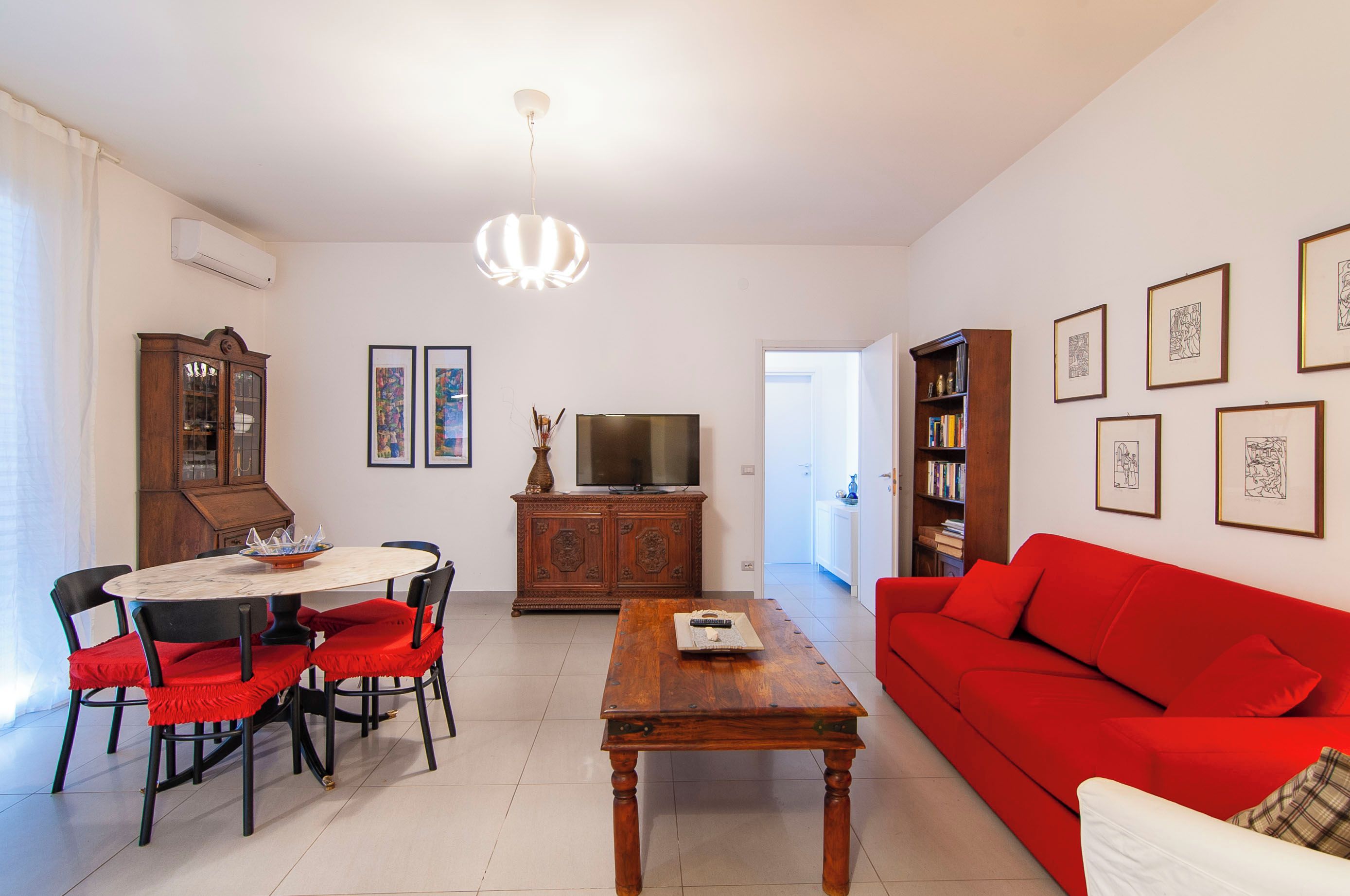 Ein Wohnzimmer mit einem roten Sofa, einem Esstisch mit Stühlen, einem Fernseher und einem Bücherregal.