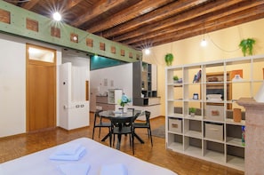 A room with a wooden ceiling, a dining table with chairs, a kitchen area, and a storage unit with shelves. - Kedimore - RomeoHouse (Verona)
