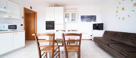 A modern kitchen with white cabinets, a microwave, and an oven. There is a dining area with wooden chairs and a table. A brown sofa is against the wall, and a flat-screen TV is on a stand.