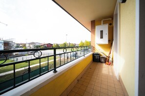 Un balcon doté d’une rambarde en métal, d’un sol carrelé et offrant une vue sur une rue bordée d’immeubles et d’arbres.