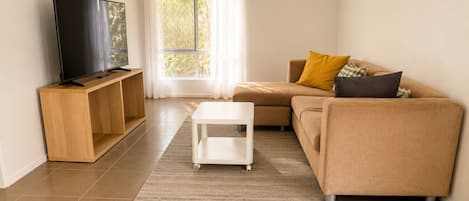 Una sala de estar con un televisor de pantalla plana sobre un soporte de madera, una mesa de centro blanca, un sofá beige con cojines y un ventanal con cortinas.