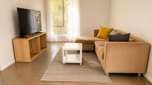 Een woonkamer met een flatscreen televisie op een houten standaard, een witte salontafel, een beige bank met kussens en een groot raam met gordijnen.