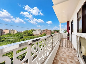 Un balcón con piso estampado, barandal blanco y vista a edificios y vegetación.