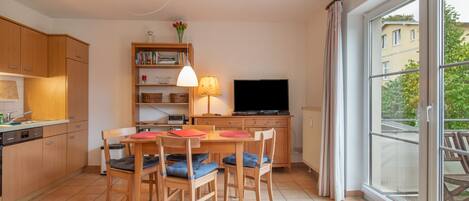 A modern kitchen with wooden cabinets, a dining table with chairs, a bookshelf, a flat-screen TV, and a large window with curtains.