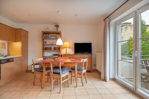 A modern kitchen with wooden cabinets, a dining table with chairs, a bookshelf, a flat-screen TV, and a large window with curtains. - Apartment 'Hafenbrise' with Sea View, Balcony, Dogs Welcome (Putbus)