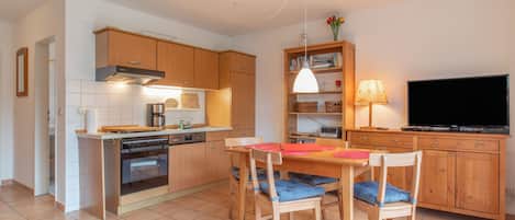 A compact kitchen with wooden cabinets, a built-in oven, a dining table with chairs, a television, and a bookshelf.