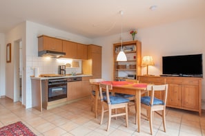 A compact kitchen with wooden cabinets, a built-in oven, a dining table with chairs, a television, and a bookshelf. - Apartment 'Hafenbrise' with Sea View, Balcony, Dogs Welcome (Putbus)