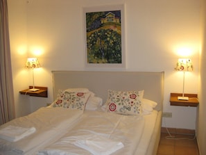 Uma cama de casal com almofadas florais, duas luminárias de cabeceira, um quadro sobre a cama e uma tomada na parede.
