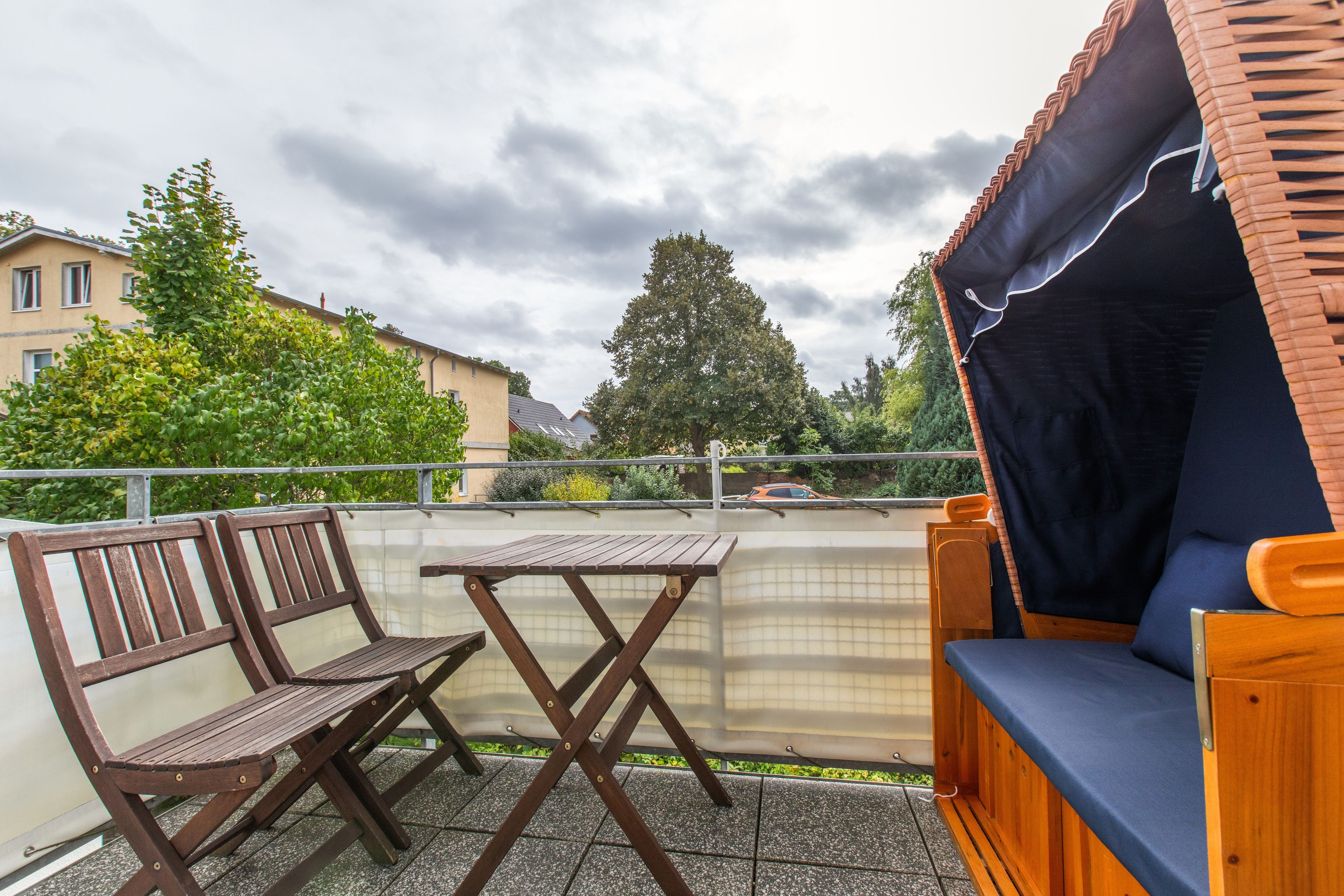Ein Balkon mit einem Holztisch und Stühlen, einer gepolsterten Bank und Blick auf die Umgebung mit Gebäuden und Bäumen.