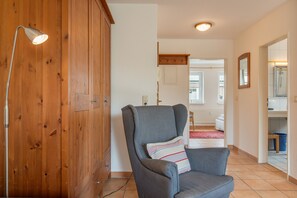 A room with a wooden wardrobe, a grey armchair with a striped pillow, a standing lamp, and a view of a bathroom with a shower and a mirror.