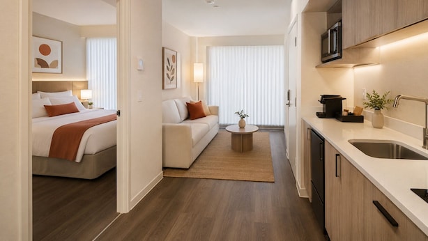 A modern hotel room with a bed, a sofa, a small round table, and a kitchen area with a sink and microwave. - Downtown Elmora Suites (Toronto)